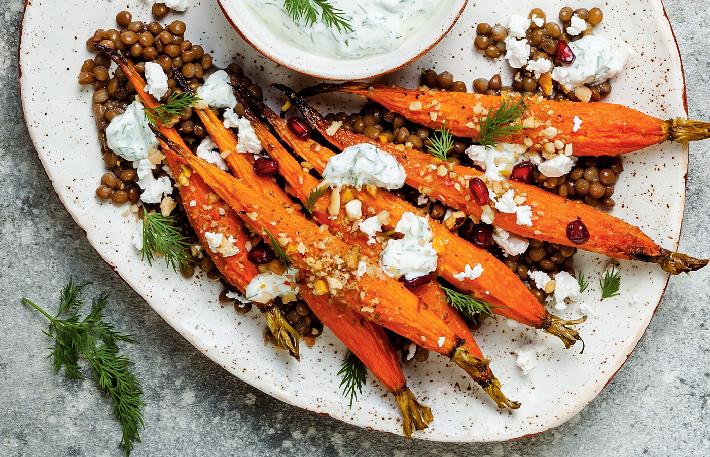 Overhead shot of carrots on a plate with dill, lentils, pomegranate seeds, and soft cheese. 
