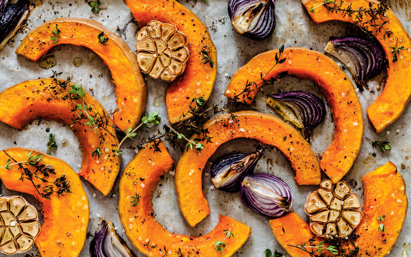 Overhead shot of sliced pumpkin, garlic, red onion, and herbs on a baking sheet.