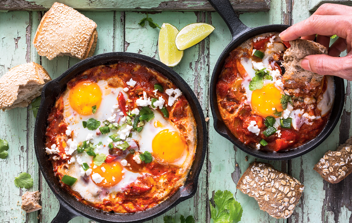 Overhead shot of shakshuka in two cast iron pans with a hand reaching from off-screen to dip crusty bread into the pan.